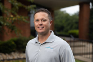 A man in a gray polo standing in front of a brick building.